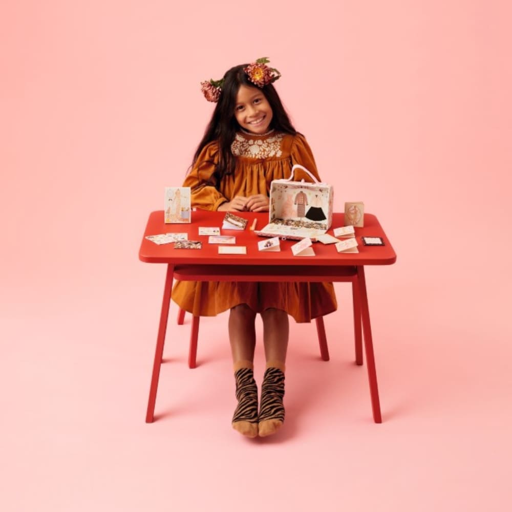 Young girl in an orange dress sitting at a red table with various items on a pink background