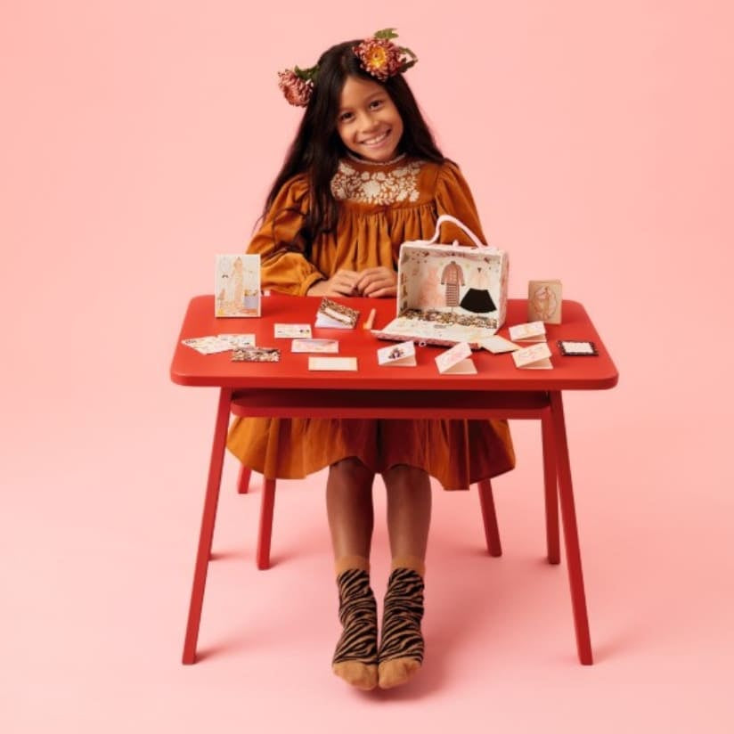 Young girl in an orange dress sitting at a red table with various items on a pink background