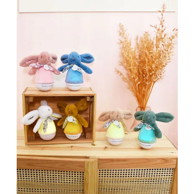 Five plush bunny toys in pastel colors displayed on and around a wooden crate and cabinet, with dried grass decor against a soft pink wall.