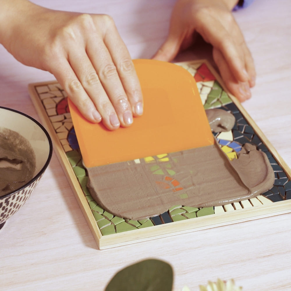 Person using a spatula to spread brown substance on a colorful mosaic board.