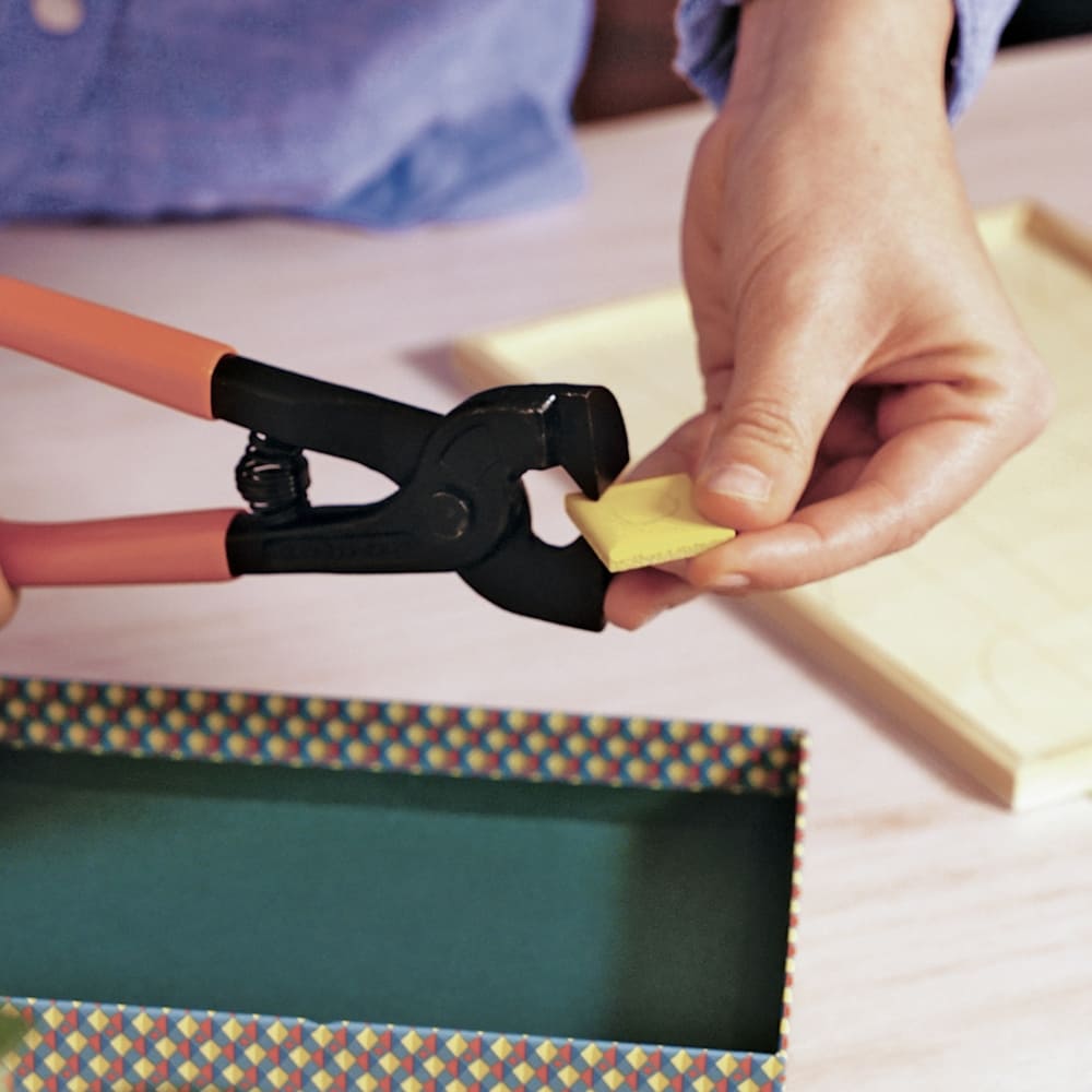 Person using a black clamp to hold a yellow object over a colorful box.
