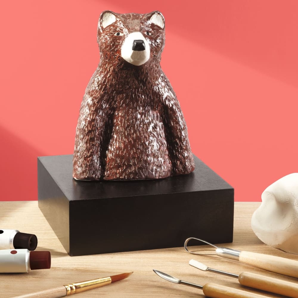 Decorative bear sculpture on a black base with art supplies on a wooden surface and pink background