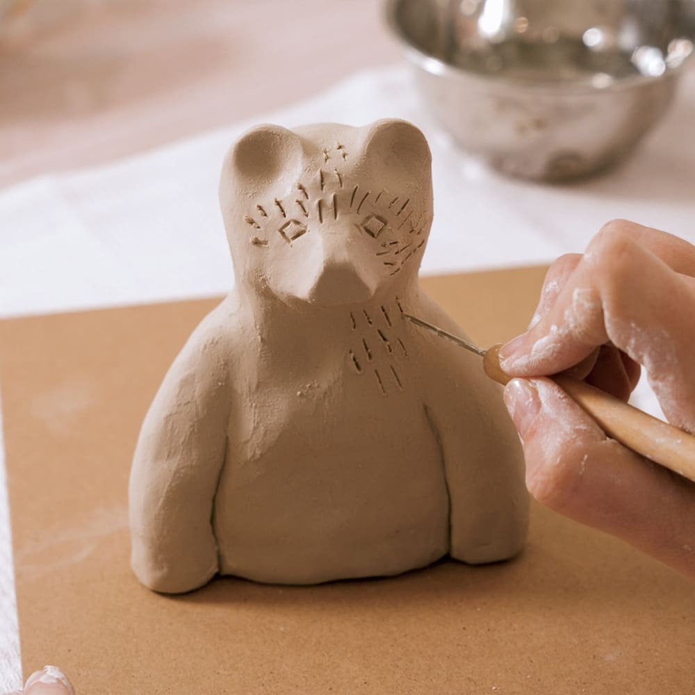 Clay sculpture of a bear being worked on with a tool on a wooden surface.