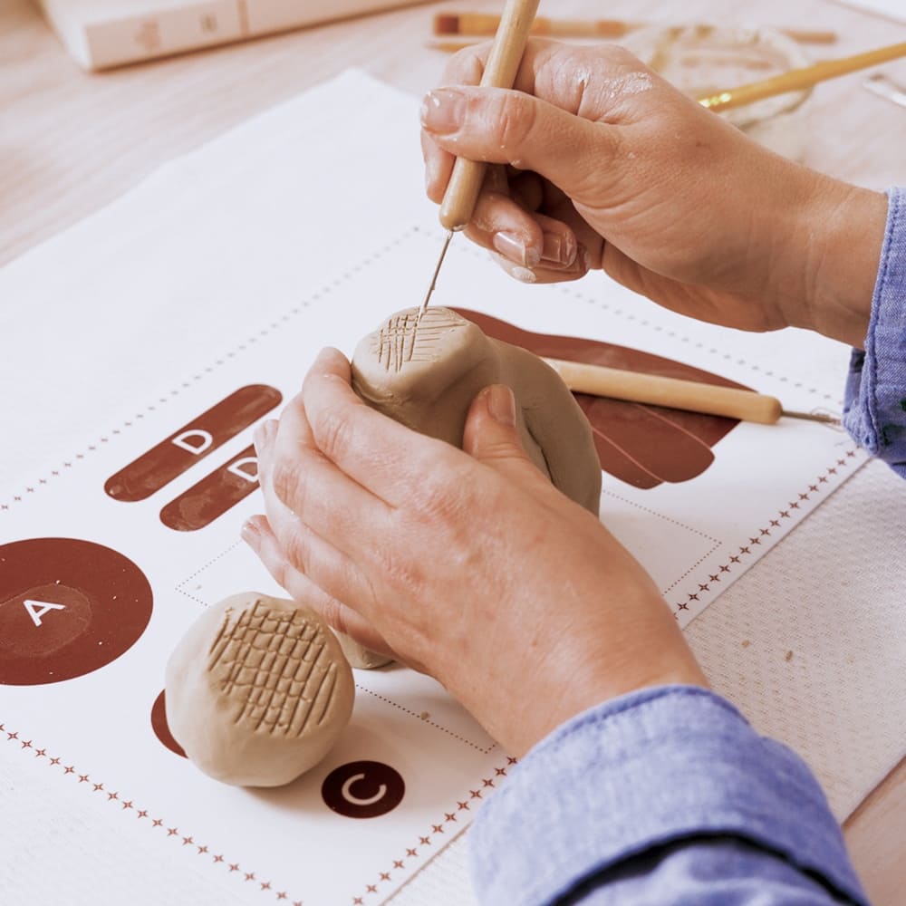 Person using a tool to texture a piece of clay on a table with a lettering guide.