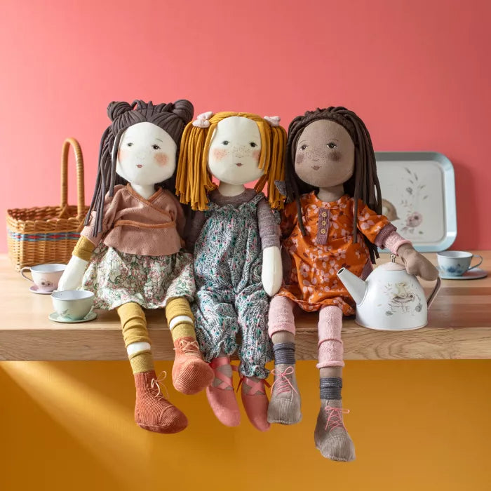 Three dolls sitting on a wooden shelf with a pink wall and yellow floor.